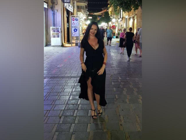 LorraineCatty, vêtue d'une robe noire avec une fente haute, des talons fins, souriante, avec des cheveux ondulés foncés, debout sur une rue pavée, le soir, des personnes en arrière-plan, seins visibles, sexy, confiante..