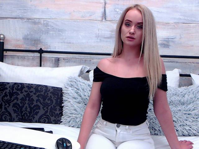 SophieXDee con i capelli biondi, una maglietta nera a spalle scoperte, pantaloni bianchi, seduta sul letto, con una testiera in metallo nero, cuscini bianchi, cuscini con motivi grigi e neri, e una tastiera nera e bianca..