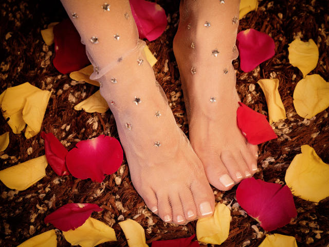PiedsMagiques' feet, adorned with star-shaped rhinestones on sheer stockings, resting on a brown, textured surface with scattered red and yellow rose petals, showing white-painted toenails.