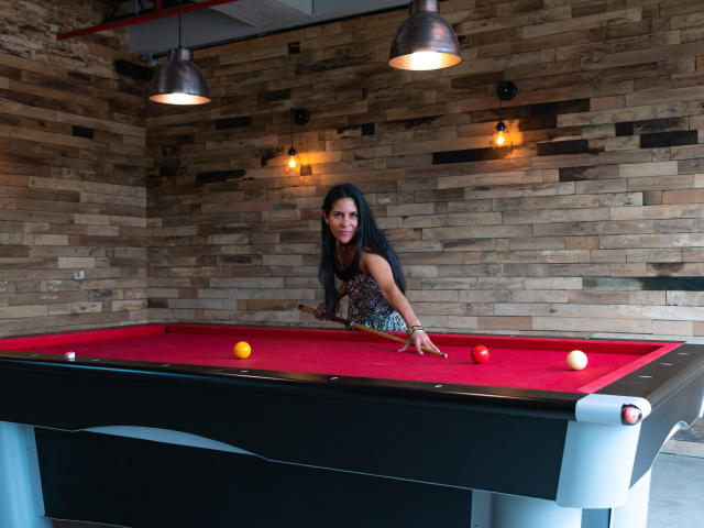 RoraimAnaggie with long black hair, wearing a sleeveless dress, leans over a red pool table, aiming a cue at a ball, with two hanging lamps above.