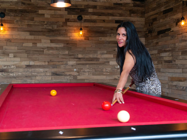 RoraimAnaggie with long black hair, wearing a leopard print dress, leans over a red pool table, holding a cue stick, with a yellow ball, red ball, and white ball.