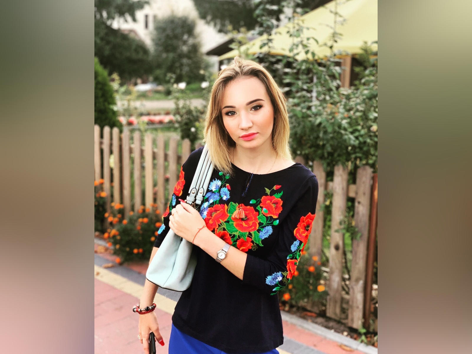 KateHailey, con cabello rubio, vestida con una blusa negra con bordados florales en rojo y azul, sosteniendo un bolso azul claro, de pie en una acera con una valla de madera..