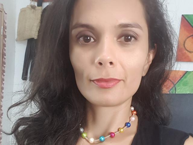 Natthally tiene una piel marrón media, cabello negro ondulado, ojos marrones y lápiz labial rosa. Lleva un collar de cuentas de colores y una blusa negra, con una sonrisa sutil..