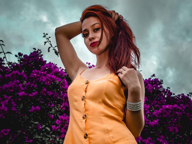 Eliza Jones, avec ses cheveux roux, portant une robe orange avec des boutons, les cheveux relevés, debout devant des fleurs violettes, avec du rouge à lèvres et un bracelet en argent..