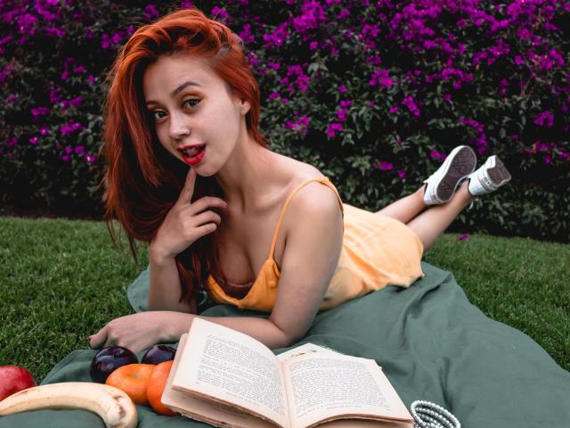 Eliza Jones, con cabello rojo, acostada en el césped con un vestido amarillo, con un dedo en la boca, un libro y fruta delante, flores moradas en el fondo, zapatillas blancas, linda, con los pechos visibles..