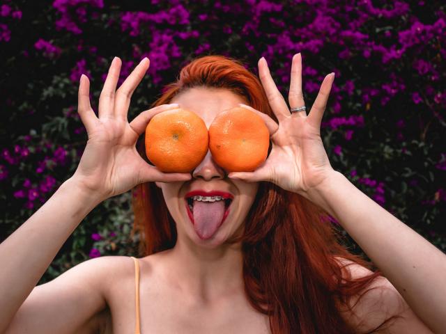 Eliza Jones, met rood haar, die twee sinaasappels voor haar ogen houdt, haar tong uitsteekt, een topje met dunne bandjes draagt, een ring om haar linkerhand, en paarse bloemen op de achtergrond..