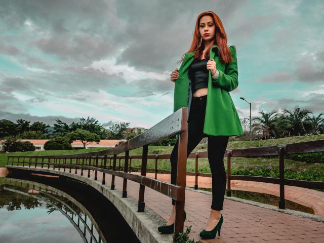 ElizaJones staat op een brug, groene jas, zwarte top, zwarte broek, groene hakken, houdt de jas vast, rood haar, bewolkte lucht, houten railing, reflecterend water, groen gras, bomen op de achtergrond..