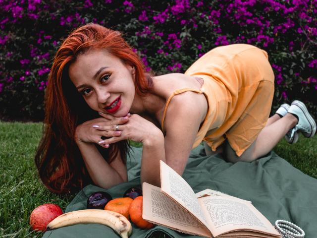 ElizaJones with red hair, orange dress, and white sneakers, on green grass, kneeling, smiling, hands on chin, book and fruit (banana, apple, orange, grapes) in front of her.