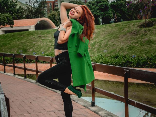 Eliza Jones, com cabelo ruivo, camiseta preta, calça jeans preta, lenço verde, salto alto verde e pulseira de prata, posando com o braço levantado, sorrindo, em pé em um caminho de tijolos, com uma perna levantada..