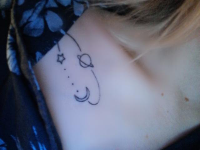 L'épaule de NaozuQAce est ornée d'un petit tatouage noir représentant un croissant de lune, une planète, une étoile et des points alignés. On peut apercevoir des cheveux blonds sur la droite..