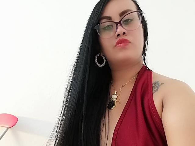 CathyCat met lang zwart haar, een rode haltertopjurk, een bril, oorringen, een ketting en rode lippenstift, met een tatoeage op haar rechter schouder zichtbaar, kijkend naar de camera..