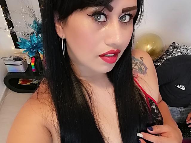 CathyCat, con i capelli neri, il rossetto rosso e gli orecchini a cerchio, mostra il seno in una maglietta nera, con un tatuaggio sulla spalla destra, seduta accanto a un ragazzo con i jeans..