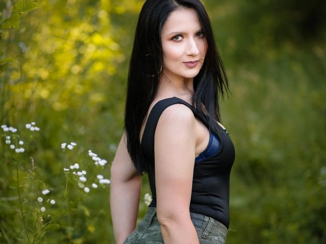 AnaHathor hat schwarzes Haar, einen blauen BH, der unter dem schwarzen Tanktop sichtbar ist, grüne Hosen, steht draußen mit weißen Blumen, lächelt, schaut über die Schulter, grüner Hintergrund, grüne Augen..