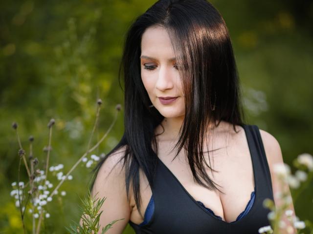 AnaHathor with black hair, looking down, wearing a black tank top showing her big tits, standing in a green, blurry outdoor setting, small white flowers in the foreground.