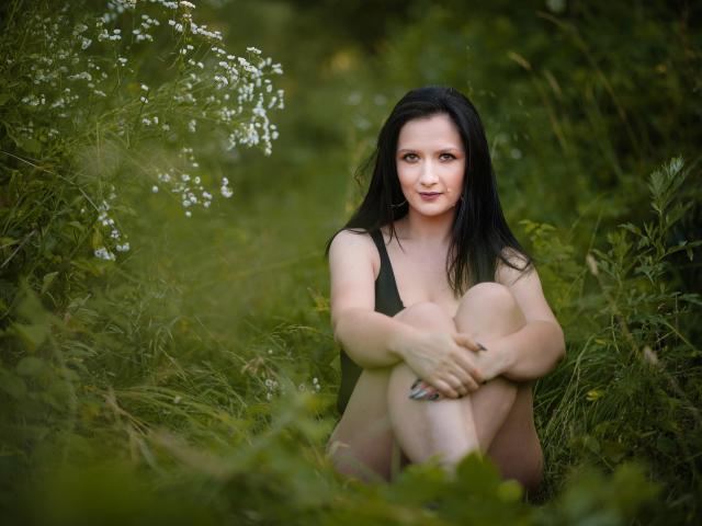 AnaHathor, sitting in green grass, has black hair, pale skin, and small tits, wearing a black top, knees up, arms around legs, smiling gently, with white flowers nearby.