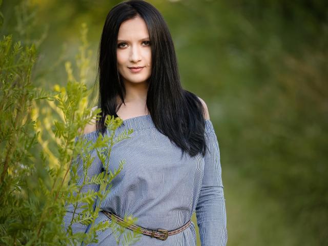 AnaHathor, avec de longs cheveux noirs, un pull bleu à une épaule, de petites seins, un "bush" vert à l'avant, debout dans un environnement verdoyant, souriant doucement, une ceinture marron, lumière naturelle, décor extérieur..