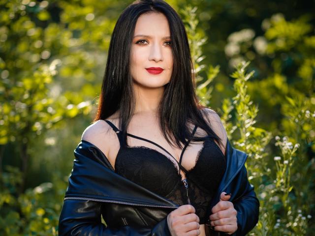 AnaHathor with long black hair, red lipstick, and black lace bra, zipping down a black leather jacket, showing her tits, standing in a sunlit green forest.