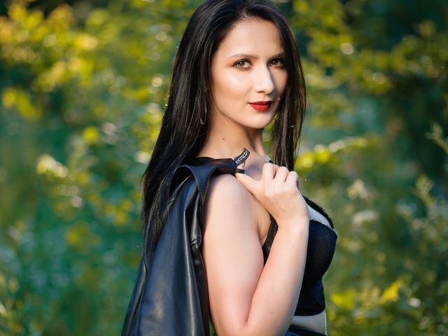 AnaHathor with long black hair, red lipstick, black leather jacket, and black top, holding jacket, standing outdoors, green blurred background, looking over shoulder, holding jacket strap.