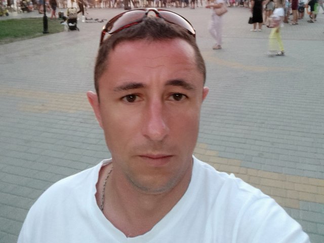 MaxyQRedhot, un homme chauve avec des cheveux bruns, portant un t-shirt blanc, a un visage légèrement rouge, et des lunettes de soleil sur la tête, avec un collier en argent, dans un endroit bondé..
