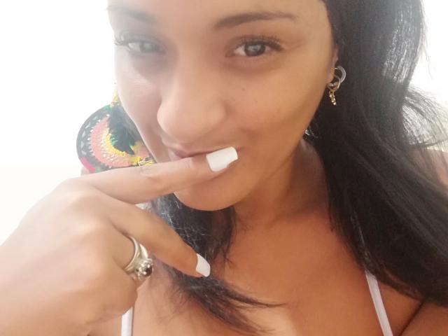 ShanelLaure, met bruine huid, lang zwart haar en witte nagels, die aan haar vinger knabbelt, draagt een witte top, gouden oorbellen en een zilveren ring, en glimlacht. Haar borsten zijn licht zichtbaar..