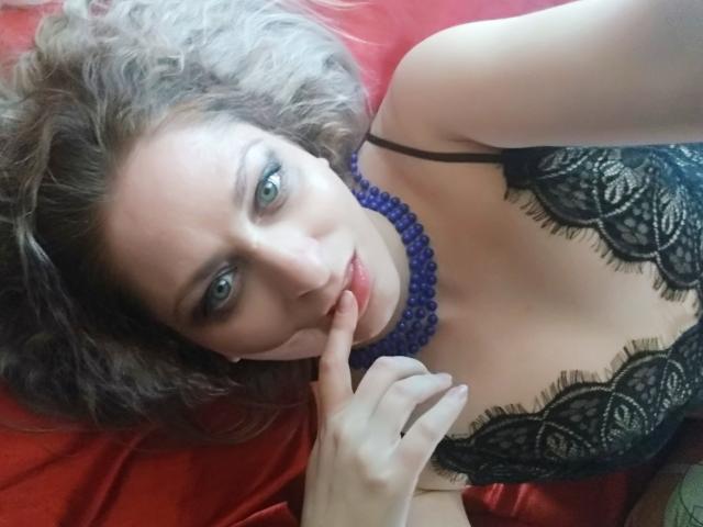MonicaMiss, con i capelli castano ricci, gli occhi azzurri e una collana di perline viola, indossa lingerie di pizzo nero, morde il dito e giace su una superficie rossa. Il seno è visibile, aspetto sexy..