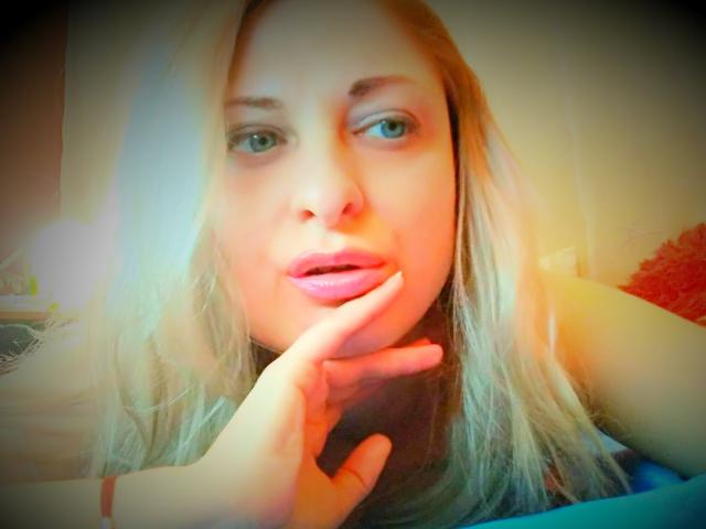 MonicaMiss with blonde hair, green eyes, and pink lips, touching her chin with a finger, looking at the camera with a slight smile. Her boobs are not visible.