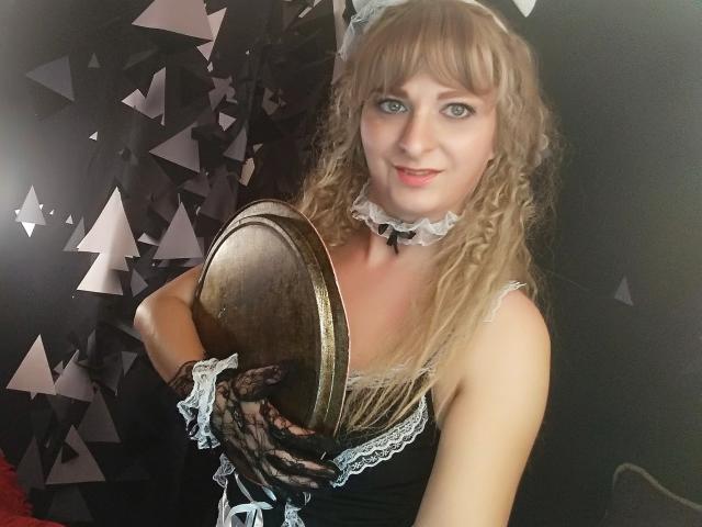 MonicaMiss mit lockigem, blondem Haar, trägt ein schwarzes Schnürcorsett, eine weiße Spitzenkette, hält eine Messingplatte und trägt schwarze Handschuhe. Sie lächelt in die Kamera und zeigt ihre Brüste..
