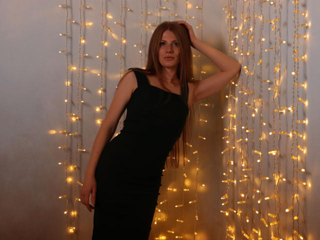 GretyArdent with long red hair, black dress, hand on head, standing against warm fairy lights, soft glow on pale skin, subtle smile, slender, sexy, confident, attractive, cute.