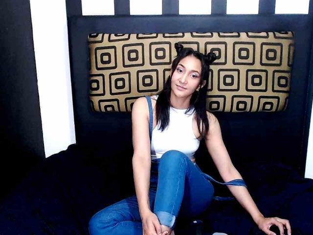 AllisonGag, avec ses cheveux noirs, portant un débardeur blanc et un jean bleu, assise sur un lit noir, les jambes croisées, et portant de petites pièces de tête en forme de cornes noires..