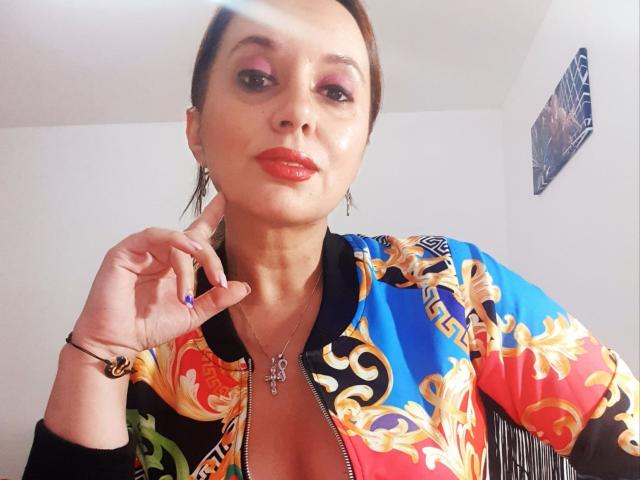 MaitresseBelle con lápiz labial rojo, sombra de ojos rosa y cabello castaño recogido en una coleta, vistiendo una chaqueta colorida con estampado floral, abierta para mostrar el escote, sosteniendo su barbilla..