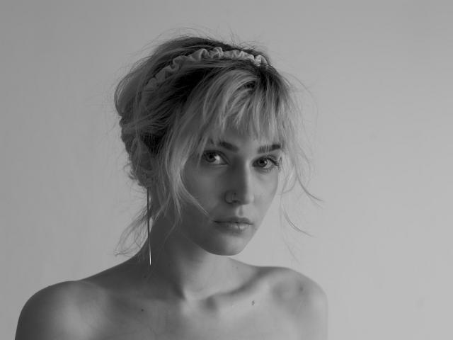 SarahMiss tiene el cabello rubio despeinado con una diadema trenzada, con una expresión seria y los pezones ligeramente visibles, fondo gris, blanco y negro, luz suave, plano medio, hombros desnudos..