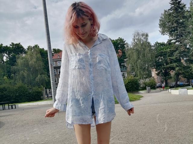 SarahMiss, avec des cheveux rose-violet, portant une chemise blanche, légèrement transparente, à boutons, debout sur un chemin de gravier, regardant vers le bas, les bras légèrement levés, dans un décor de parc..
