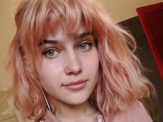 SarahMiss a les cheveux roses et ondulés, des yeux verts et un anneau de nez. Elle porte une chemise rose avec des boutons blancs et regarde l'appareil photo avec un léger sourire..