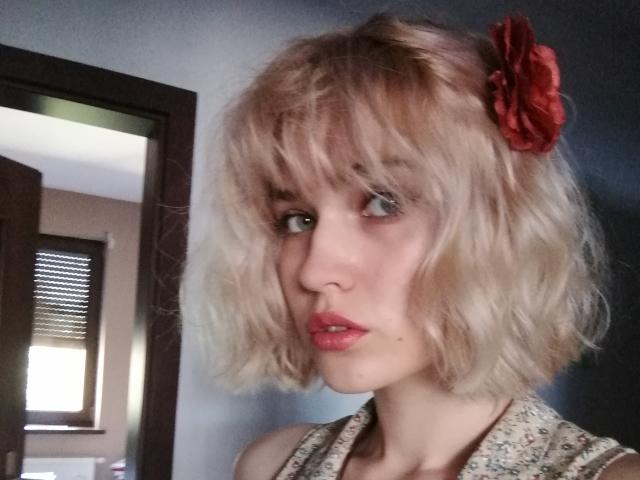 SarahMiss heeft blond, golvend haar dat tot op haar schouders reikt, met een rode bloem, bleke huid, blauwe ogen en roze lippenstift, en draagt een top met bloemen, met een licht geïrriteerde uitdrukking..
