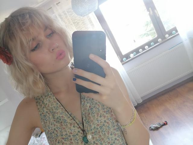 SarahMiss, com cabelo loiro ondulado, piercing no nariz e vestido floral, fazendo beicinho em frente ao espelho, segurando um celular, chão de madeira, pulseira e um pequeno carrinho de brinquedo à direita..