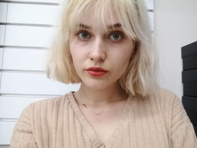SarahMiss has blonde, wavy hair, green eyes, red lips, a nose ring, and wears a beige, ribbed sweater. She's looking straight at the camera with a neutral expression.