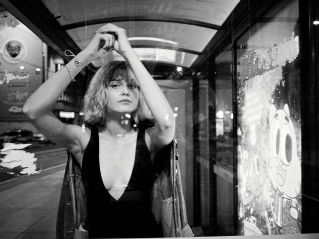 Photo en noir et blanc de SarahMiss, avec des cheveux blonds courts, les bras levés, portant une robe à col en V profond qui révèle ses petites seins, à l'intérieur d'une station d'autobus en verre, de nuit..