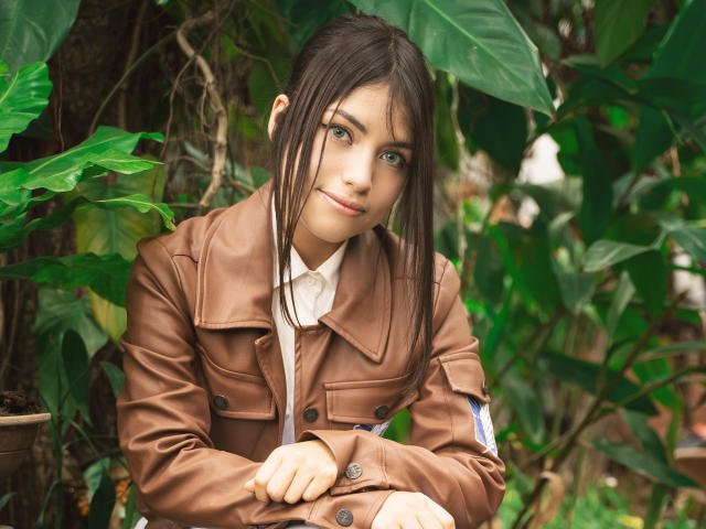 VanityValos has green eyes, dark brown hair in pigtails, wearing a brown leather jacket, white shirt, and sitting with arms crossed, surrounded by green leaves.