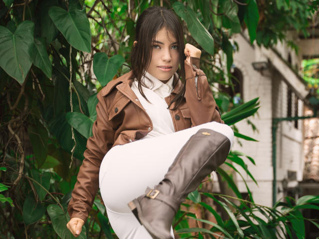 VanityValos with brown jacket, white pants, and brown boots, sitting in green leafy background, one leg raised, hand touching brown jacket, showing her tits and confident, sexy smile.