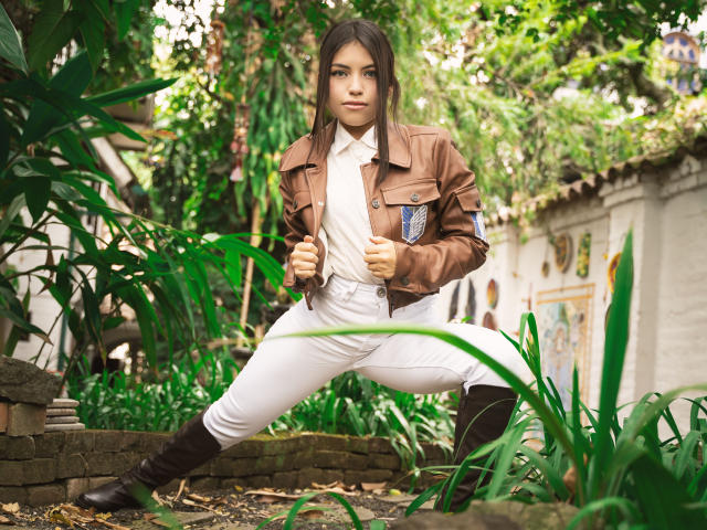 VanityValos, met een bruin jasje, witte broek en bruine laarzen, hurkt in een groene, bladerrijke tuin, vuisten gebald, met een vastberaden blik, zichtbare bruine ogen en recht, donker haar..