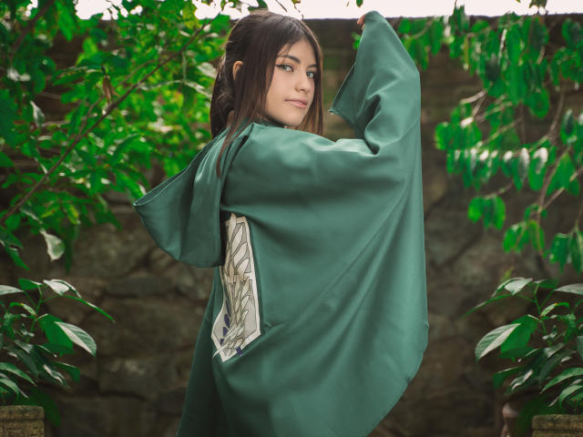 VanityValos con veste verde, braccia alzate, che guarda oltre la spalla, capelli castani, occhi verdi, seno piccolo, sorriso appena accennato, circondata da foglie verdi, parete rocciosa sullo sfondo..