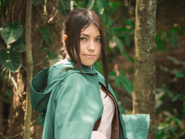 VanityValos mit grünem Umhang, braunem Hemd und süßem Gesicht, steht im Wald. Sie hat dunkelbraunes Haar, grüne Augen und ein leichtes Lächeln und blickt über ihre Schulter..