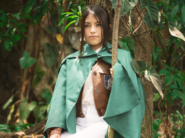 VanityValos with green cape, brown leather vest, white pants, holding tree branch, standing in jungle, green leaves, brown tree bark, serious expression, brown hair, blue eyes, medium tits.