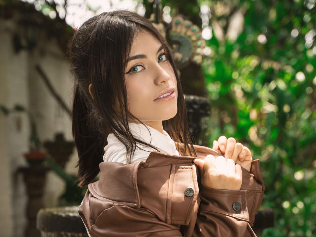 VanityValos with brown hair, green eyes, and a cute face, wearing a brown jacket, white shirt, and looking over her shoulder, holding her jacket. Her tits are partially visible.