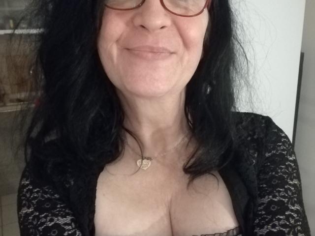 Emilianne with black wavy hair, glasses, smiling, showing cleavage in a black lace top, heart necklace, fair skin, medium-sized boobs.