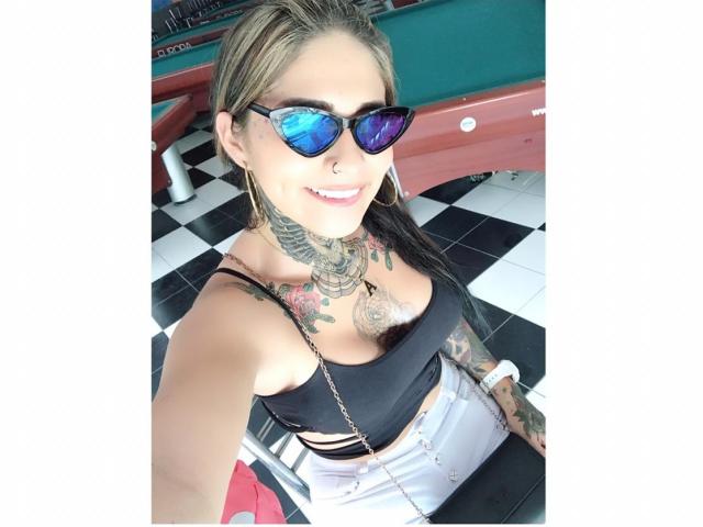 CristalHugeX, avec des tatouages, portant un débardeur noir, un pantalon blanc taille haute et des lunettes de soleil bleues en forme de cœur, souriant à l'appareil photo dans une pièce lumineuse..