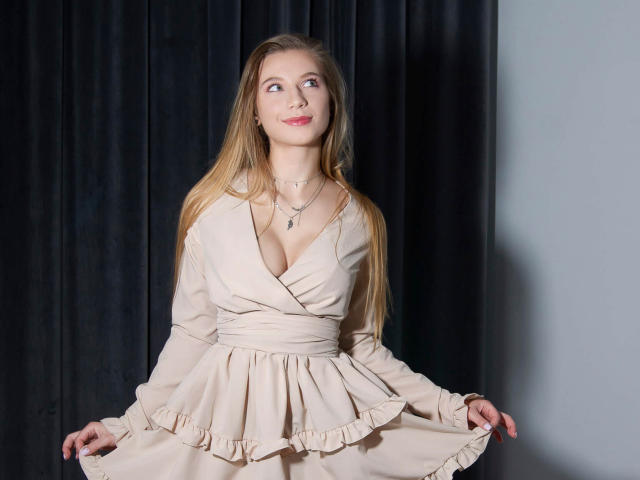 LucyBestCandy with long blonde hair, smiling, wearing a beige dress with a deep V-neck, showing her boobs, holding the skirt, necklace, standing against dark curtains.