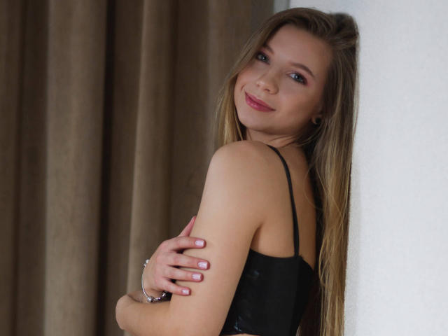 LucyBestCandy mit langen blonden Haaren, schwarzem Spitzenoberteil, verschränkten Armen, lächelnd in die Kamera, rosafarbenen Nägeln, weichem Licht, weißer Wand, braunem Vorhang, entspannt, sexy, dezentes Make-up..