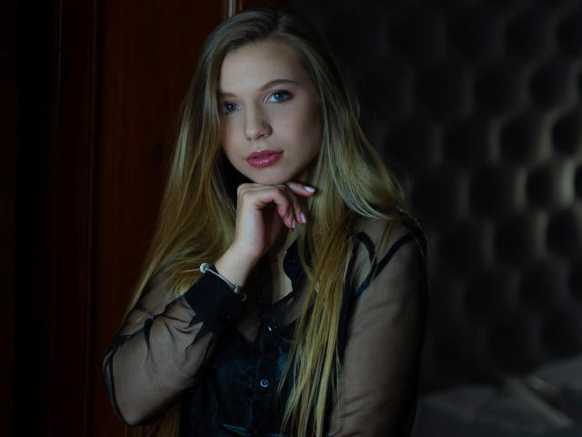 LucyBestCandy mit blonden Haaren, blauen Augen und rosafarbenen Lippen, trägt ein durchsichtiges schwarzes Oberteil und legt ihr Kinn in die Hand, wobei sie ihre Brüste und ein verführerisches Lächeln zur Schau stellt..