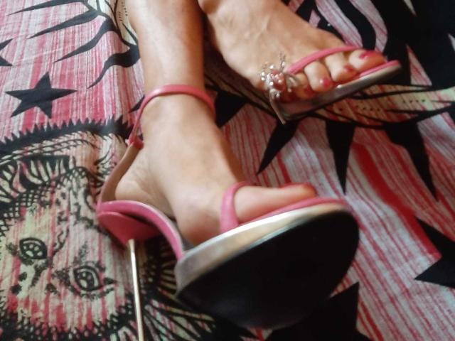 Los pies de PinkyTs, con sandalias rosas de tacón alto con tachuelas plateadas en los dedos, tobillos cruzados, mostrando uñas de los pies pintadas de rosa, sobre una tela estampada..